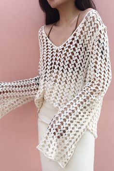 a woman wearing a white top with crochet on the shoulders and sleeves, standing against a pink wall