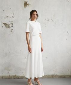 a woman standing in front of a wall wearing a white dress with short sleeves and an asymmetrical neckline