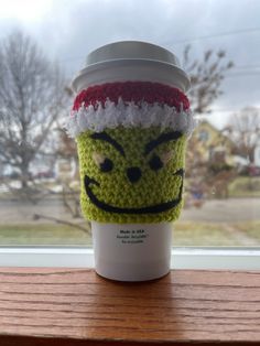 a coffee cup with a knitted grin face on it's lid sitting on a window sill