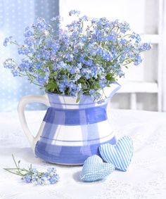 blue flowers in a pitcher on a table with two heart shaped eggs next to it