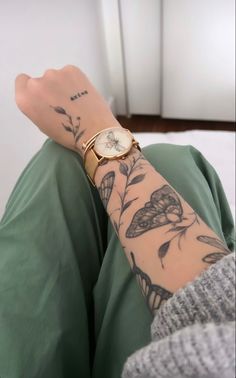 a woman's arm with tattoos and a watch on her left wrist, sitting in front of a white wall