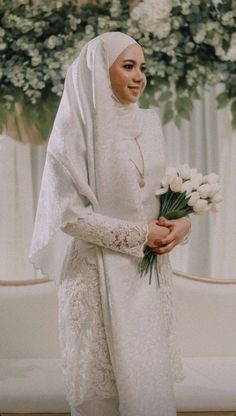 a woman in a white dress holding flowers and smiling at the camera while wearing a headscarf