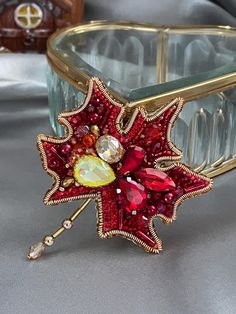 a red and gold brooch sitting on top of a silver table next to a glass container