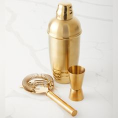 a gold cocktail shaker and two brass cups on a white marble counter with a golden snake handle