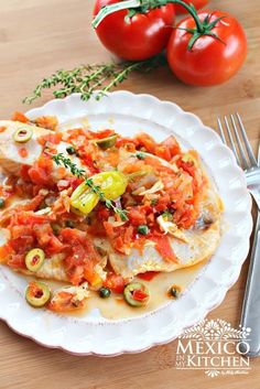 a white plate topped with fish covered in tomatoes and olives next to a fork