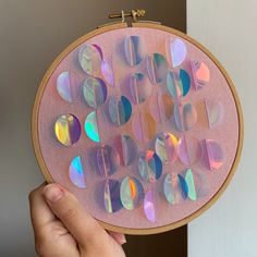 a hand holding up a pink embroidery with many different colored circles on it and a wooden hoop hanging from the wall