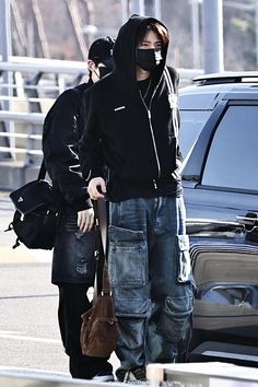 a man in black hoodie standing next to a parked car and holding a brown bag