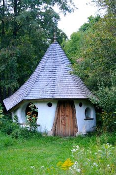 a small white building with a wooden door in the middle of some grass and trees