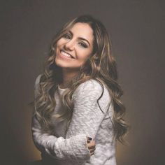 a woman with long hair smiling and wearing a white sweater, standing in front of a dark background