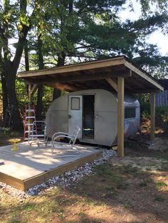 a small white trailer sitting in the middle of a forest next to a shed and trees