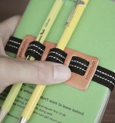 a hand is holding two pencils in front of a green book with black and white stitching