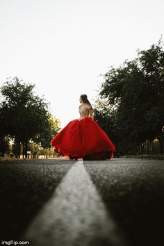 a woman in a long red dress is walking down the street with trees behind her