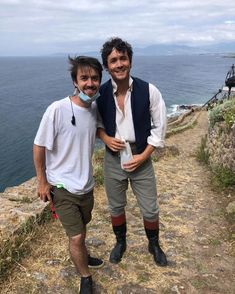 two men standing next to each other on top of a hill near the ocean,