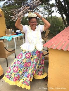 a woman in a colorful skirt and hat is holding branches above her head while sitting at an outdoor table