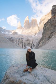 a woman sitting on top of a rock next to a body of water with mountains in the background