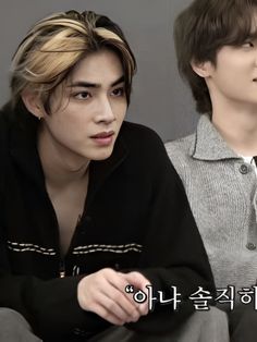 two young men sitting next to each other in front of a gray wall with korean writing on it