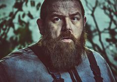 a man with a long beard and suspenders stares at the camera while standing in front of trees