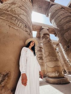 a woman in a white dress and straw hat standing next to large stone pillars at the egyptian temple