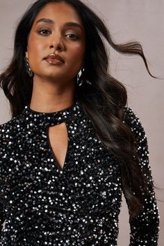 a woman with long hair wearing a black sequin dress and large hoop earrings, standing in front of a gray background