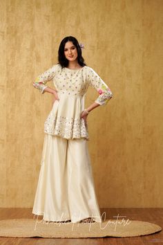 "Introducing our stunning Ivory Peplum Top with Sharara Pant, adorned with mesmerizing mirror and resham embroidery, crafted from luxurious Chanderi silk fabric." The Peplum Top is exquisitely designed to flatter the silhouette, with a chic and contemporary look. The intricate mirror and resham embroidery on the top adds a touch of elegance, creating a stunning visual effect. The top features a round neckline, complemented with a sleeveless design, which makes it perfect for any special occasion Peplum With Skirt Indian, Peplone Design, Unstitched Off White Kurta With Mirror Work, Chanderi Palazzo Set With Gota Work For Reception, Off White Chanderi Sets With Mirror Work, Navratri Raw Silk Sharara With Chikankari Embroidery, Designer Off White Kurta With Mirror Work, Bollywood Style Chanderi Palazzo Set In Off White, Off White Palazzo Set With Zari Work For Festivals