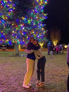 two people hugging each other in front of a large christmas tree with lights on it