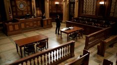 an empty courtroom with wooden benches and tables in it's center, as a man stands at the desk