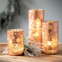 three copper colored candles sitting on top of a wooden table next to a pine tree