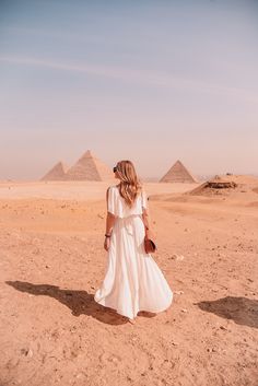 a woman in a white dress is walking through the desert with pyramids behind her