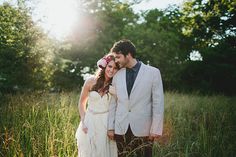 a bride and groom are standing in tall grass with the sun shining through the trees behind them