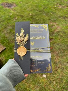 a person holding up a black and gold wedding card with flowers on the front cover