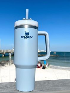 a travel mug sitting on top of a wooden table next to the ocean and beach