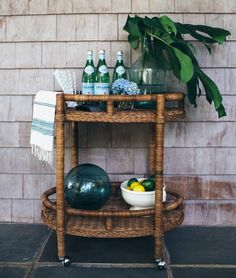 a wicker bar cart with bottles and bowls on the top, next to a potted plant