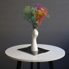 a white vase with colorful flowers sitting on top of a small table in front of a gray wall