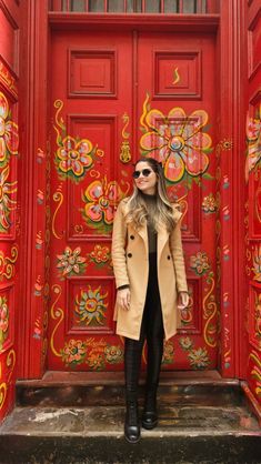 a woman standing in front of a red door wearing black boots and a tan coat