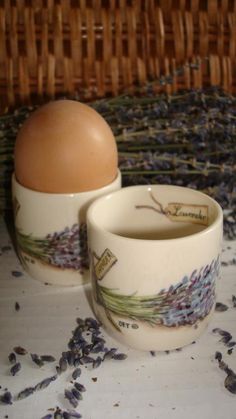 two cups and an egg sitting on a table with lavenders scattered around the bowls