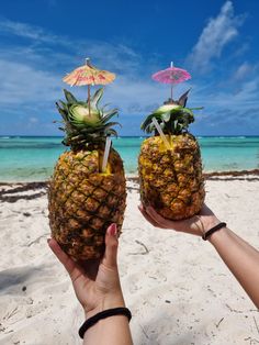 two pineapples on the beach with umbrellas sticking out of them, one being held up by someone's hand