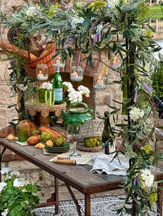 a table topped with lots of food and greenery next to a stone wall covered in flowers