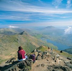 two people sitting on the top of a mountain looking down at some water and mountains