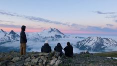 three people sitting on top of a mountain looking at the clouds and mountains in the distance