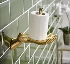 a toilet paper holder with two gold hands holding a roll of toilet paper in front of a green tiled wall