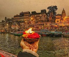 a person holding a bowl with flowers on it in front of a river filled with boats