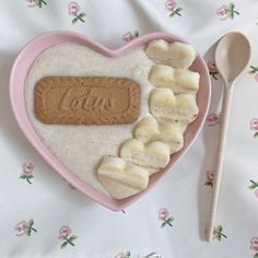 a pink heart shaped bowl filled with cereal and banana slices next to a spoon on a floral tablecloth