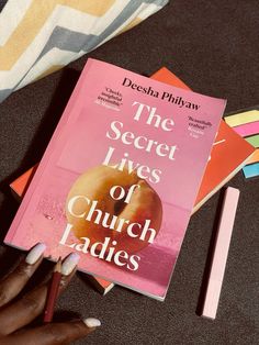 a woman's hand holding a pencil next to a pink book with the secret lives of church ladies written on it