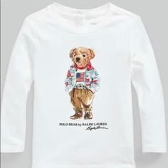 Polo Ralph Lauren For Kids Long Sleeve Top From Polo Ralph Lauren Cute Polo Ralph Lauren Long Sleeve Top In White With A Brown Teddy Bear With Cool Clothes Printed On The Front. "Polo Bear By Ralph Lauren" Is Printed In Black Letters Below The Bear. The Polo Ralph Lauren Top Has Long Sleeves, And It Has A Narrow Ribbed Edge In The Neckline. White Tops With Character Print For Fall, Long Sleeve Cotton Shirt With Character Print, Long Sleeve Cotton Top With Cartoon Print, Graphic Tee Shirt With Cartoon Print, Long Sleeve Graphic Tee With Character Print, Playful White Long Sleeve T-shirt, Polo Bear Ralph Lauren Logo Striped, Ralph Lauren Bear Sweater, Long Sleeve Cotton T-shirt With Cartoon Print