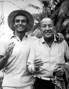 two men standing next to each other in front of some trees and palm trees with one man wearing a straw hat