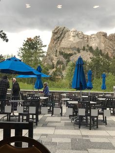 tables and chairs with blue umbrellas in front of a rocky mountain range on a cloudy day