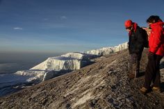 two people standing on the side of a snow covered mountain looking at something in the distance