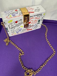 a small white box with gold chains and a chain around it on a purple surface