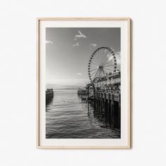 a black and white photo of a ferris wheel on the water with clouds in the background