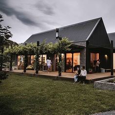 two people sitting on the porch of a black house with green grass and trees around it